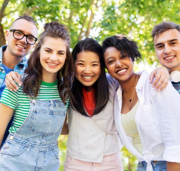 A diverse group of five young friends smiling outdoors, standing closely together under leafy trees on a sunny day, conveying joy and friendship discussing about the difference in British and American spellings