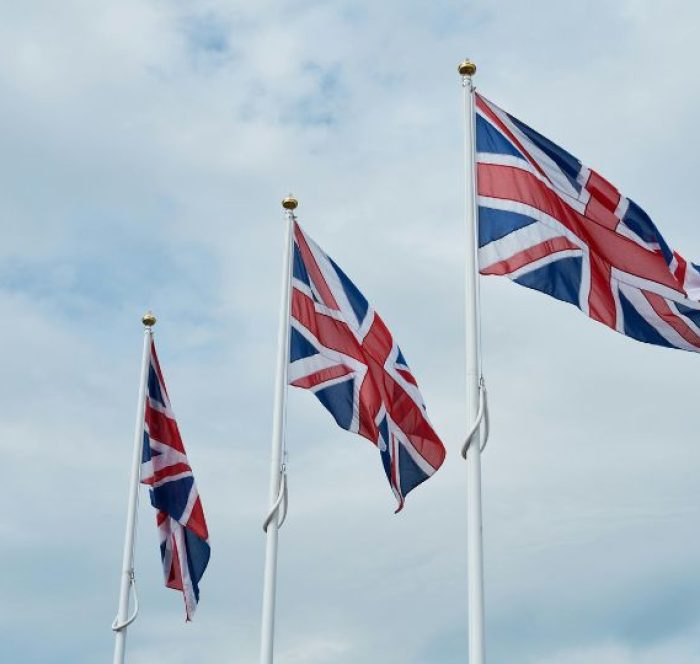 Three UK flags on white poles wave against a cloudy sky, conveying a sense of national pride and unity. The flags are evenly spaced in a row.