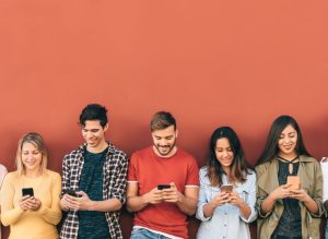 A diverse group of five young adults stand against a red wall, smiling as they look at their smartphones and checking about the apps for learning English. The mood is cheerful and connected.