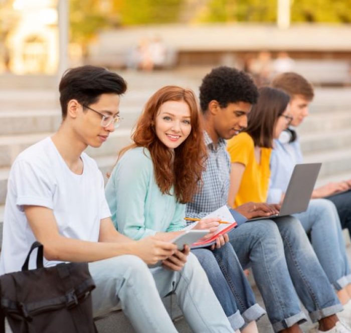 A diverse group of young adults sit on outdoor steps with laptops and notebooks, engaged in study about either and neither. One smiles at the camera, creating a friendly atmosphere.