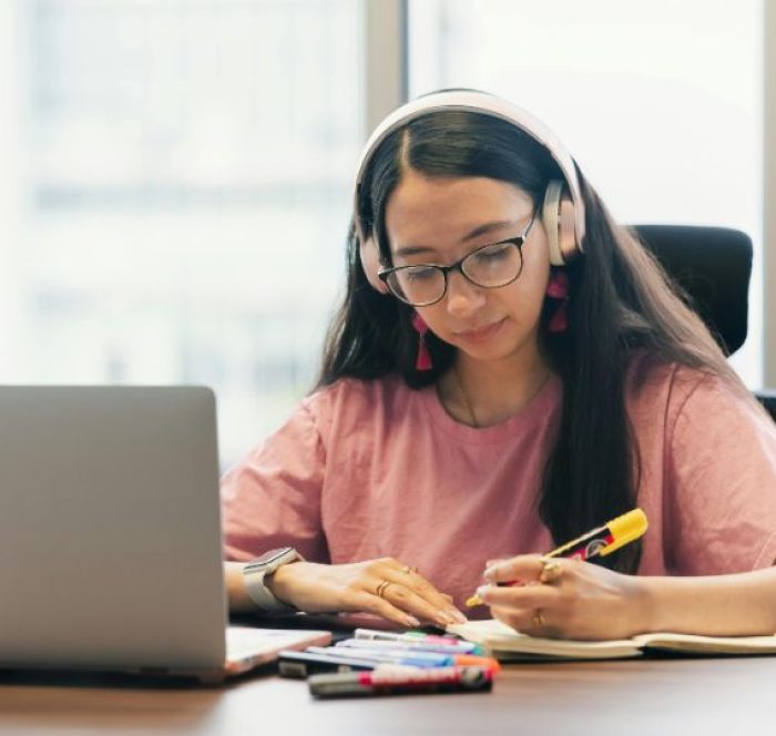 Young woman in a pink shirt and headphones focuses on writing in a notebook at a desk with a laptop and colorful markers, conveying concentration.