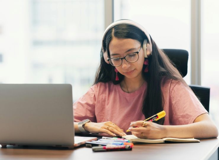 Young woman in a pink shirt and headphones focuses on writing in a notebook at a desk with a laptop and colorful markers, conveying concentration.