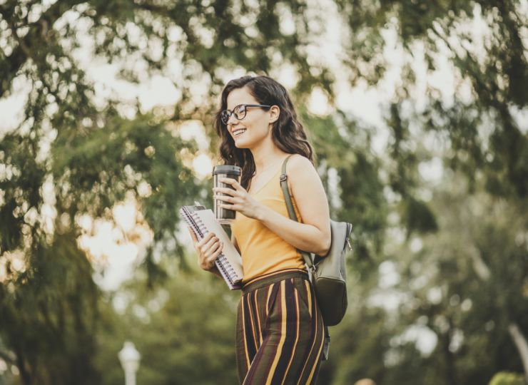 A smiling woman with glasses walks outdoors, holding a notebook and a coffee cup. She's wearing a yellow top and striped pants, surrounded by trees.