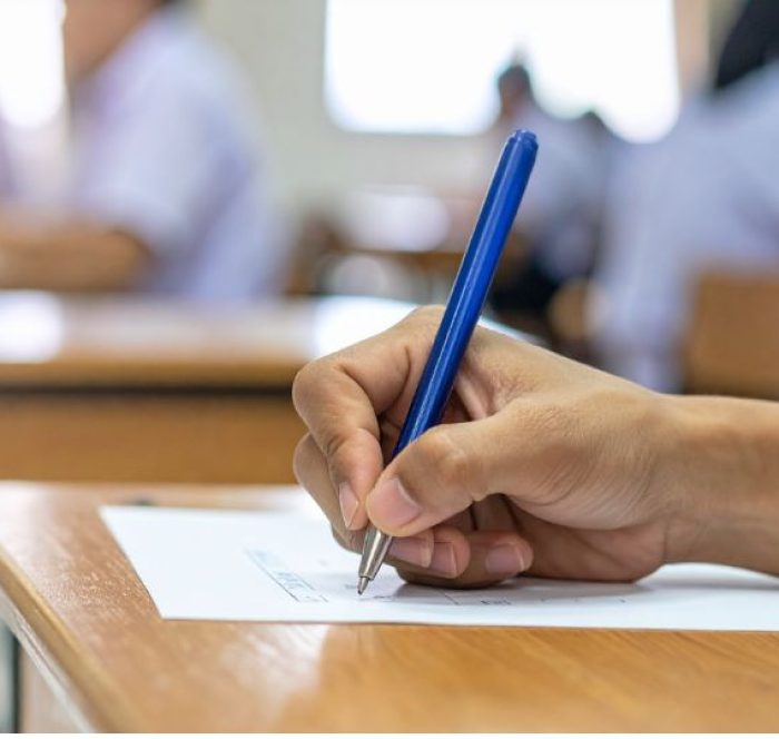 A hand holding a blue pen writes on paper atop a wooden desk in a bright classroom. Student taking test.