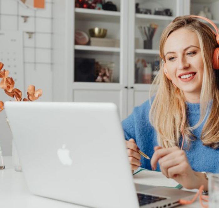 A woman in blue sweater, wearing headphones, smiles while looking at a laptop. A notepad and glass are on the table, with shelves and a calendar behind her.