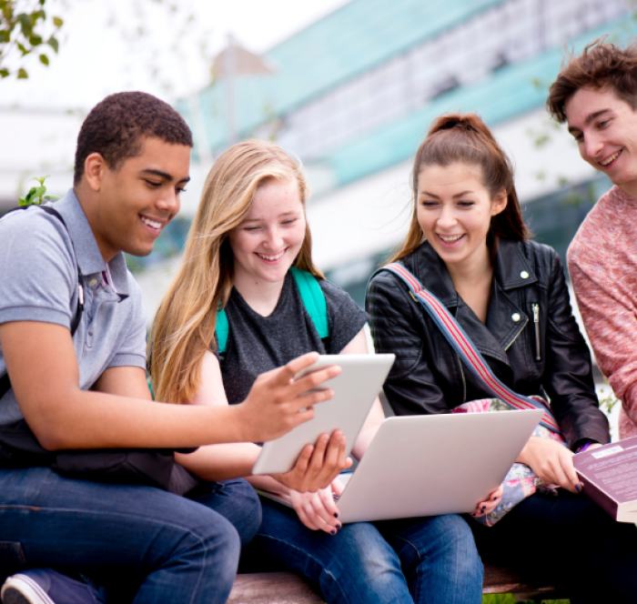 A group of four young adults, smiling and engaged, sit outdoors. They gather around a tablet and laptop, suggesting a collaborative, cheerful atmosphere.