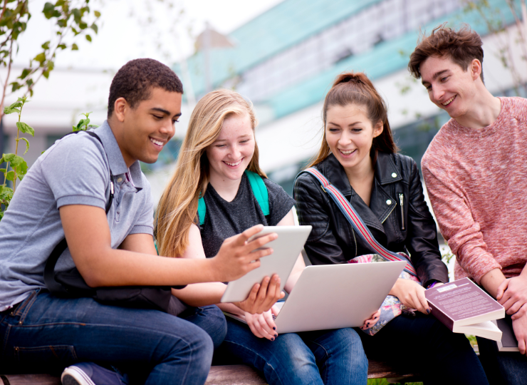 A group of four young adults, smiling and engaged, sit outdoors. They gather around a tablet and laptop, suggesting a collaborative, cheerful atmosphere.