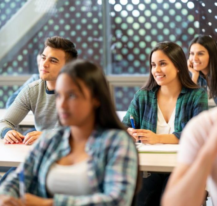 Students sit attentively in a classroom, focusing on someone ahead. They appear engaged and cheerful. A patterned window wall is visible behind them.