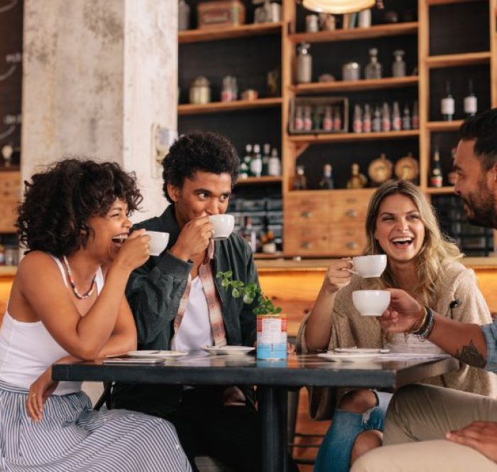 Four friends enjoying coffee at a cozy café. They are laughing and chatting, creating a warm and joyful atmosphere. Shelves with jars behind them.