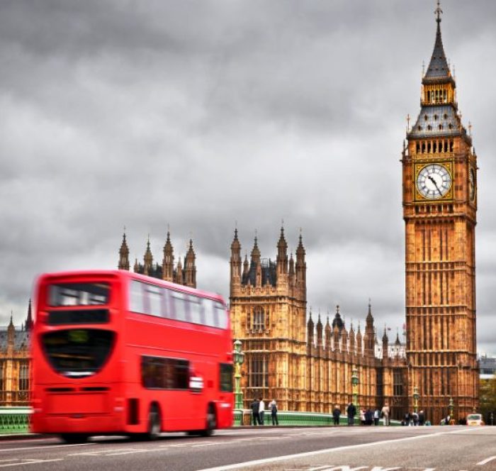 A vibrant red double-decker bus crosses a bridge in front of the iconic Big Ben and the Houses of Parliament under a cloudy gray sky.