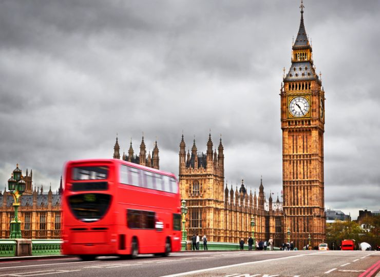 A vibrant red double-decker bus crosses a bridge in front of the iconic Big Ben and the Houses of Parliament under a cloudy gray sky.