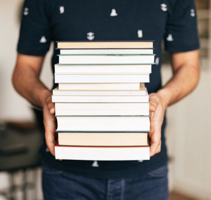 A person in a navy shirt holds a large stack of variously colored hardcover books. The scene exudes a sense of knowledge and academic dedication.