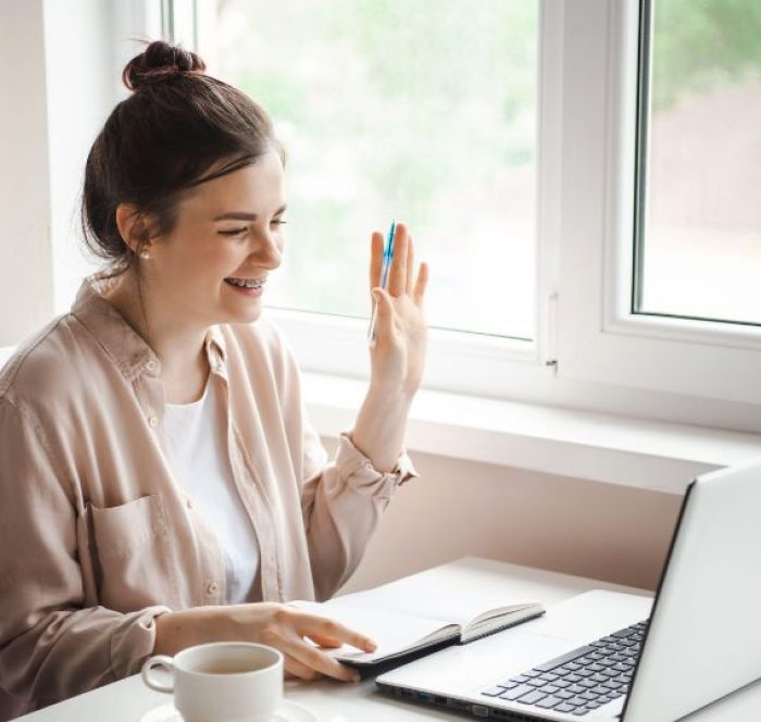 Young woman with a bun smiling and waving at a laptop screen during a video call. She holds a pen, with an open notebook and writing about 10 ways to say yes or no in English