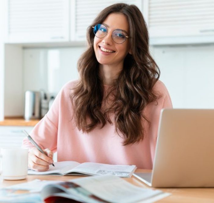 Smiling woman with long brown hair and glasses, wearing a pink sweater, writes in a notebook at a desk with an open laptop and cup, in a bright kitchen regarding the Business English Words for Work & Careers