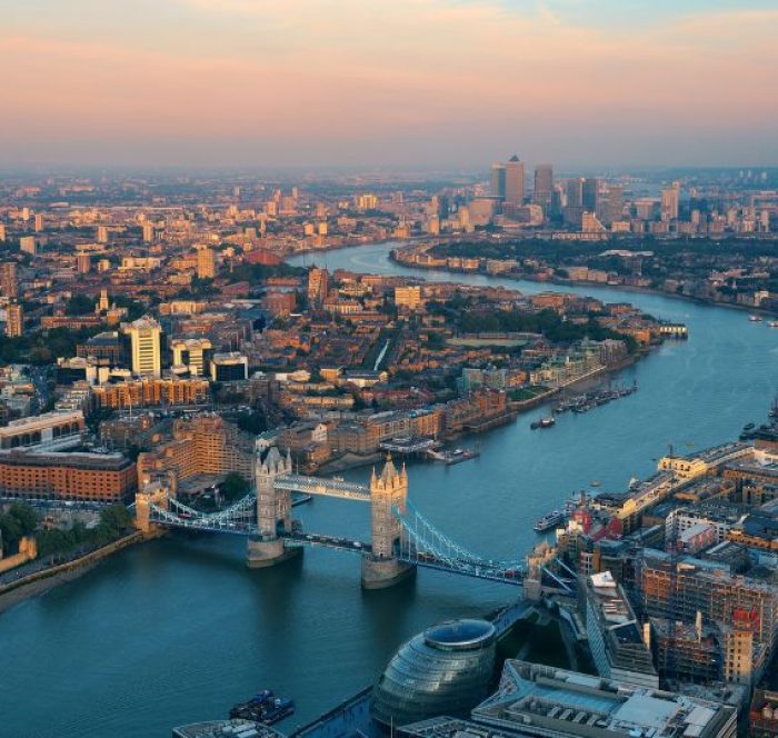 Aerial view of London at sunset, featuring Tower Bridge spanning the Thames River. Skyscrapers rise in the distance under a pink and orange sky.