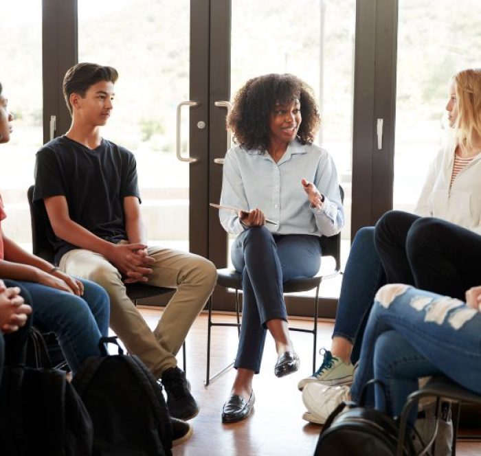 A diverse group of teenagers sits in a circle during a discussion. A woman in the middle gestures while speaking, fostering an open, friendly atmosphere.