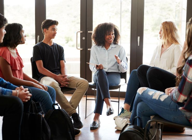 A diverse group of teenagers sits in a circle during a discussion. A woman in the middle gestures while speaking, fostering an open, friendly atmosphere.