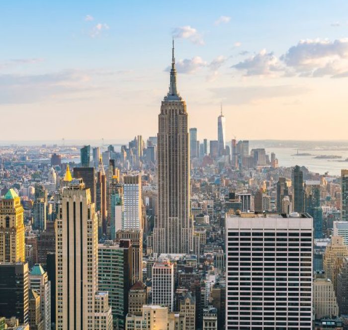 Aerial view of New York City with the Empire State Building in the center. Skyscrapers surround it under a partly cloudy, serene sky at sunset.