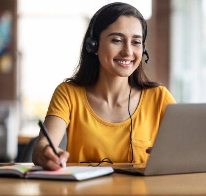 Smiling woman in a yellow shirt, wearing headphones, writes in a notebook while using a laptop regarding the IELTS TEST. Bright, cheerful room with blurred background.