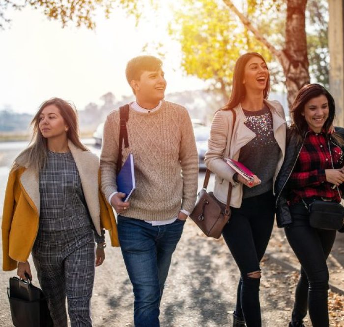 Four young adults walk along a sunny autumn street, smiling and chatting. They wear casual clothes and carry books, exuding happiness and camaraderie.