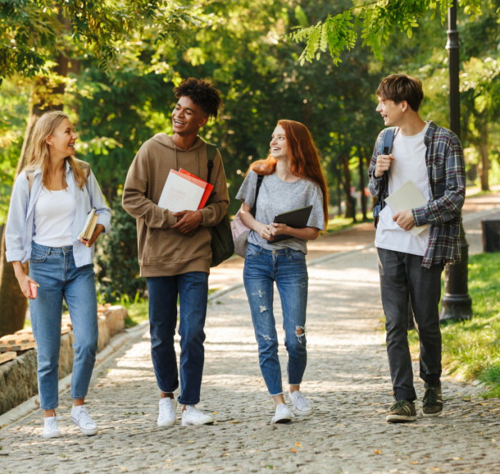 Four college students walk on a tree-lined path, laughing and carrying books. The scene is sunny, evoking a cheerful and relaxed atmosphere.