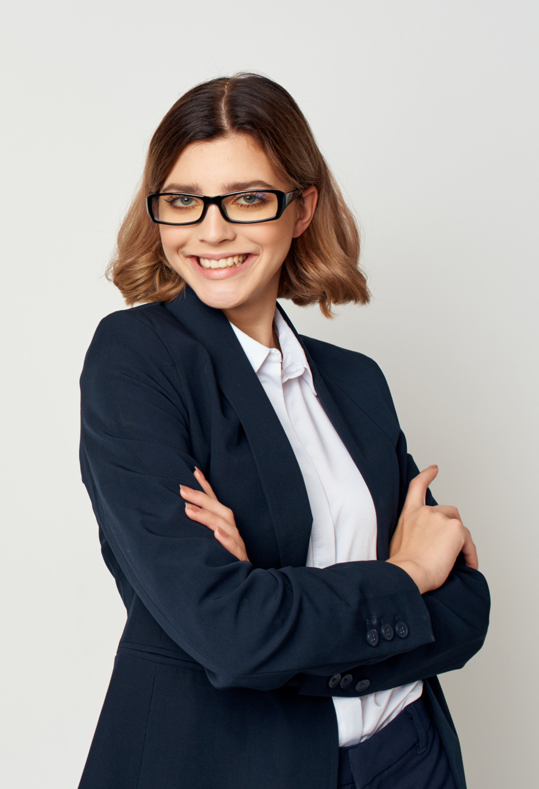 A confident woman in a navy blazer and glasses, smiling with arms crossed against a plain background, conveying professionalism and approachability.