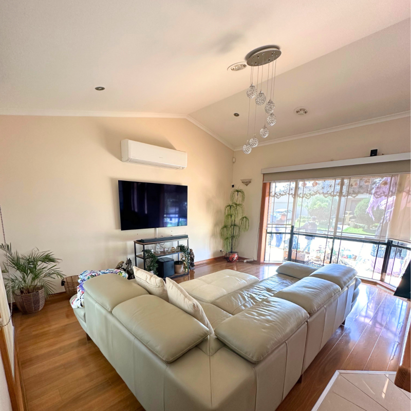 Spacious living room with a large beige sectional sofa facing a wall-mounted TV. Floor-to-ceiling windows fill the room with natural light. Elegant decor.