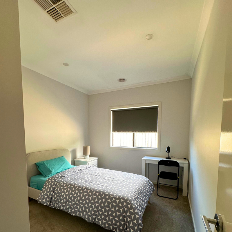 A clean bedroom with beige walls features a bed with a geometric gray duvet and turquoise pillow, a nightstand with a lamp, a desk, chair, and a window with a blind.