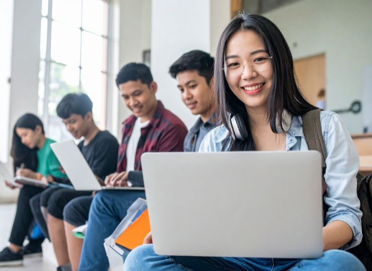 A group of diverse students sit indoors, using laptops. A happy young woman in the foreground smiles at the camera, conveying a cheerful, studious atmosphere.