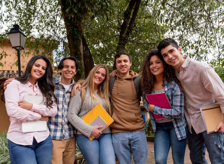 A group of six young adults stands closely together, smiling warmly, and holding books and notebooks. They're outdoors in a park-like setting, conveying a cheerful and friendly atmosphere.