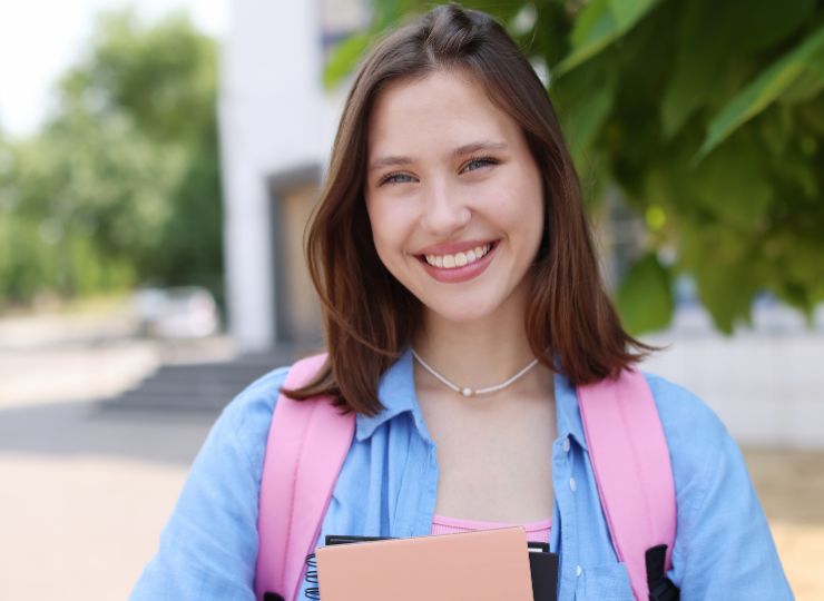 Smiling young woman with brown hair, wearing a pink backpack and holding notebooks, stands outdoors near greenery, exuding a cheerful and confident vibe.