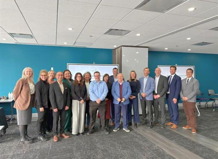 A diverse group of fifteen people, in formal attire, stand smiling in a conference room with blue walls and a screen, conveying a professional atmosphere.