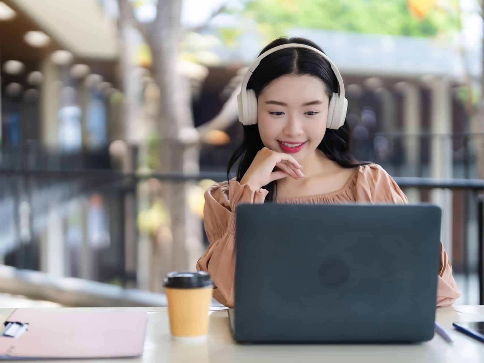 A woman in headphones smiles at a laptop in an outdoor cafe. She is relaxed and engaged. A coffee cup and notebook are on the table.