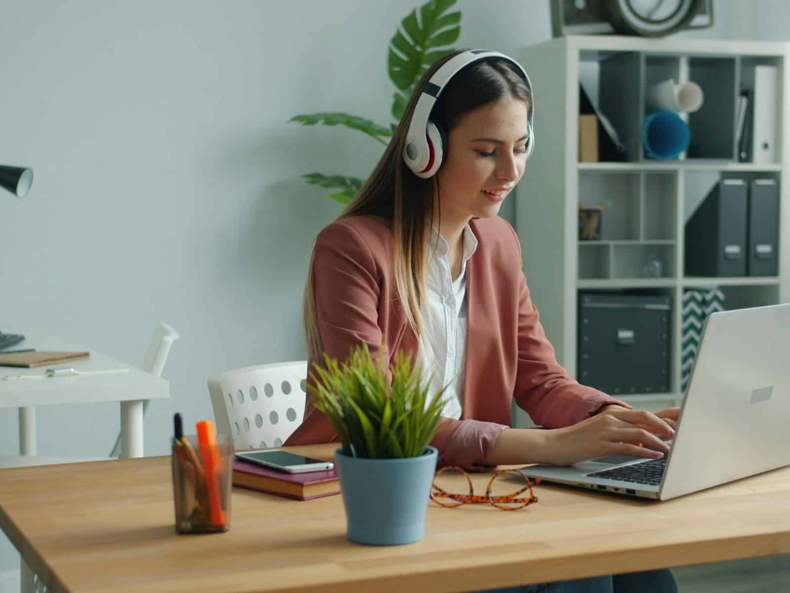 Woman in a pink blazer with headphones, typing on a laptop at a desk with potted plant, books, and stationery, conveys focus and productivity.