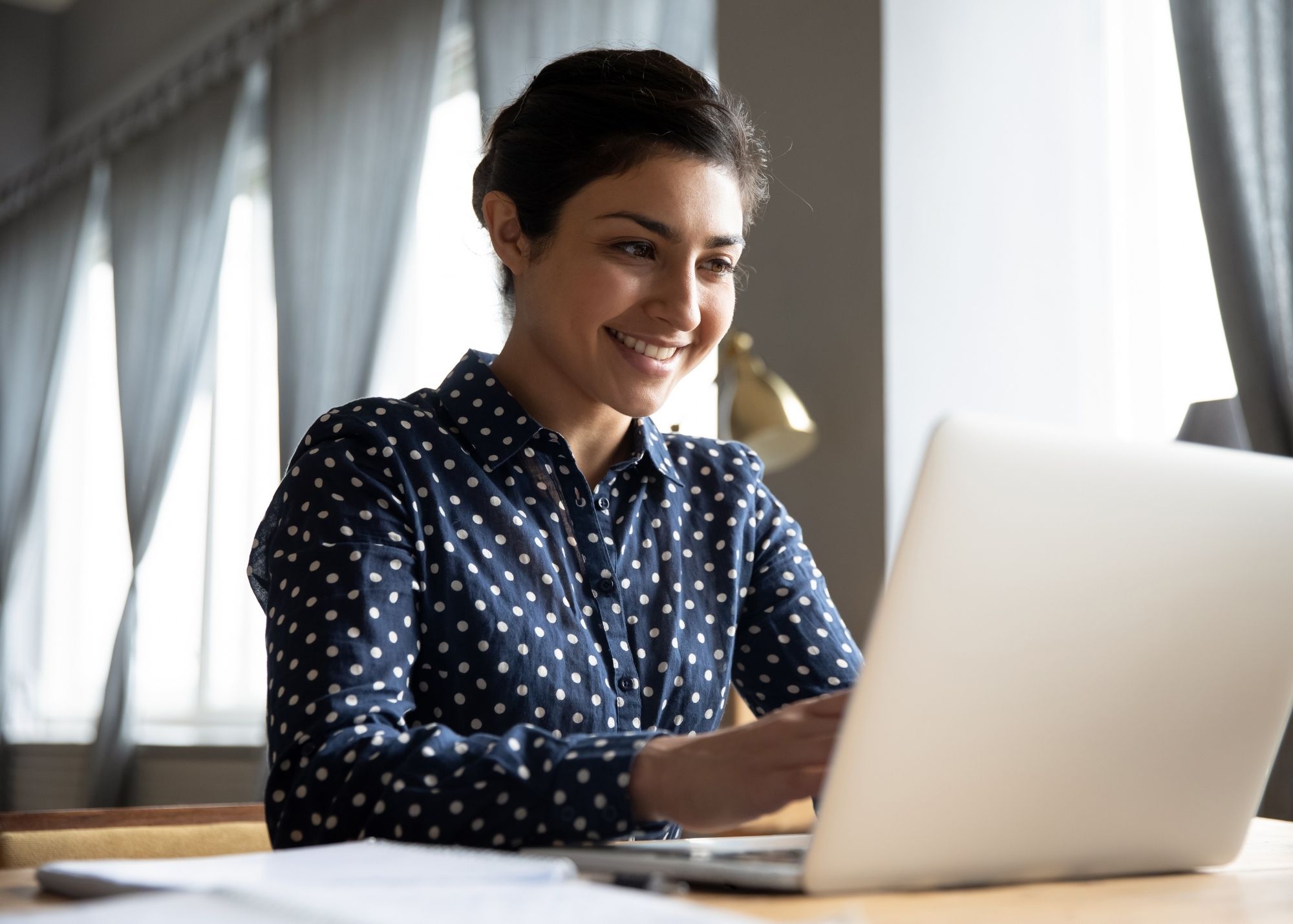 Smiling woman in a polka dot shirt working on a laptop at a wooden desk, with soft natural light coming through sheer curtains in the background.